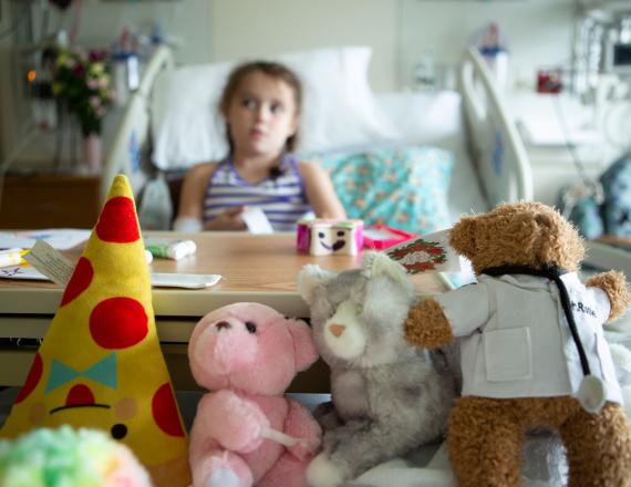 A young girl reclines in a hospital bed with four stuffed animals at her feet.