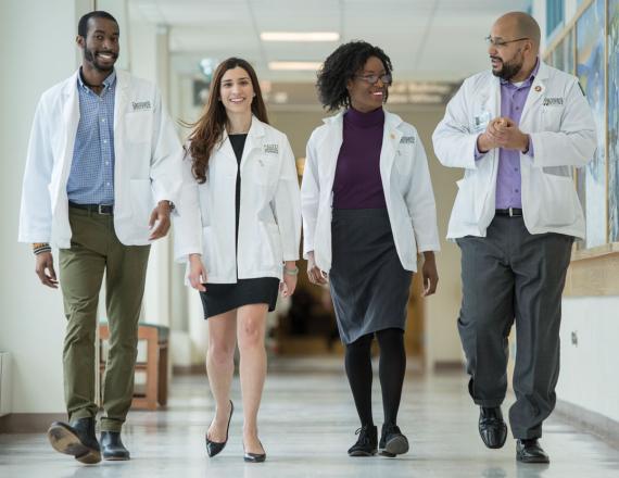 Four medical students wearing their white coats walk down the hall of a hospital.