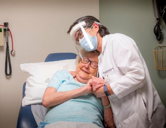 An older patient, reclining in a chair, smiles gratefully as she’s embraced by her nurse.