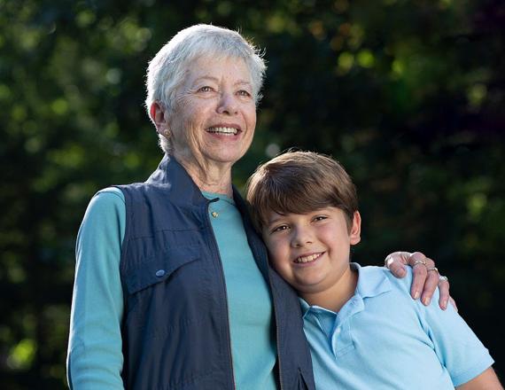 An older woman stands with her arm around her young grandson, both of them smiling happily.