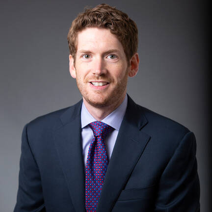 A formal headshot portrait of Dr. Linton Evans against a dark neutral studio background. He is smiling, with short brownish hair and a neat beard, dressed in a navy suit jacket, a light blue shirt, and a red and navy patterned tie.