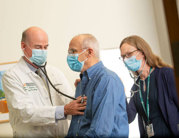 Two caregivers with a patient, one applying a stethoscope to the patient’s chest, the other to his back.