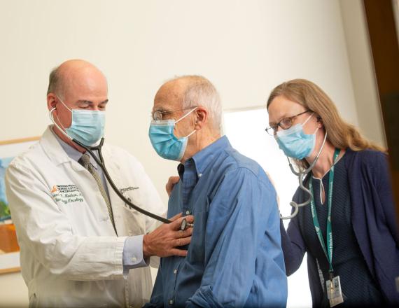 Two caregivers with a patient, one applying a stethoscope to the patient’s chest, the other to his back.