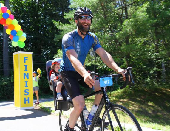 A man riding a bicycle with a young child in a baby seat behind him. They’ve just crossed a finish line.