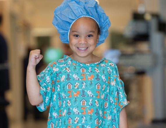 A little girl in a hospital gown and hair cap pumps her fist and smiles.