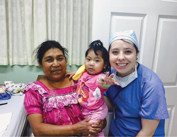 A young woman in scrubs has her arm around another woman who’s holding a little girl. The girl reaches out toward the camera.