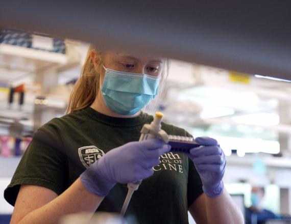 A young woman in a Geisel School of Medicine t-shirt examines the contents of a test tube rack in a laboratory.