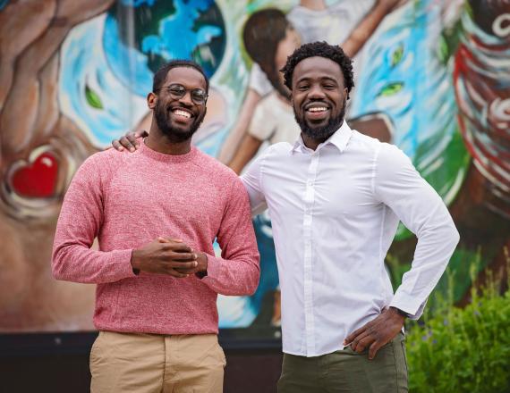 Two young men, both Geisel students, stand smiling in front of a colorful mural.