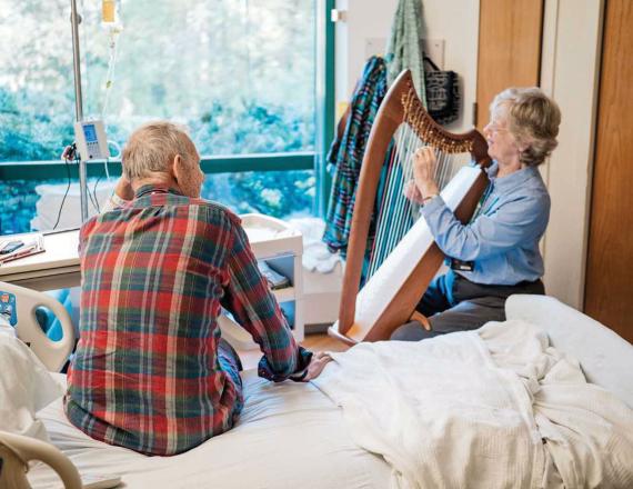 A harpist plays for a patient who’s sitting up on his hospital bed.