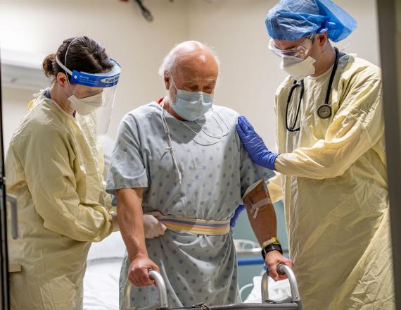 An older patient in a hospital gown grasps a walker while supported by two caregivers.