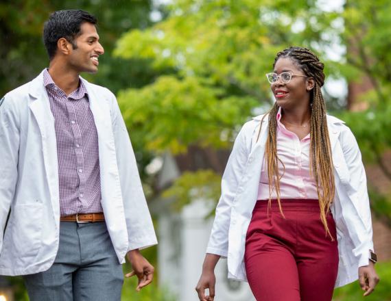 A young man and a young woman, Geisel medical students, walk outside in their white coats.