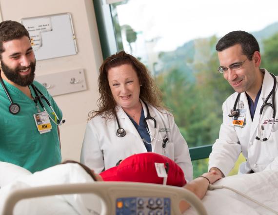 Three providers stand beside a hospital bed. One of them holds the patient’s hand.