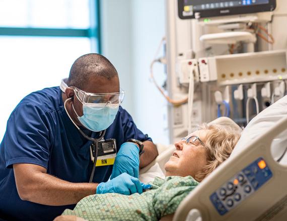 A nurse bends over a patient in a hospital bed, listening to her chest with a stethoscope.