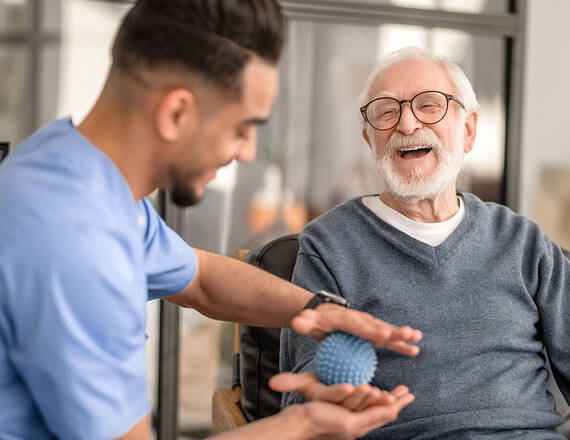 A male healthcare provider assists an elderly man with physical therapy for his hand; they are smiling and laughing.