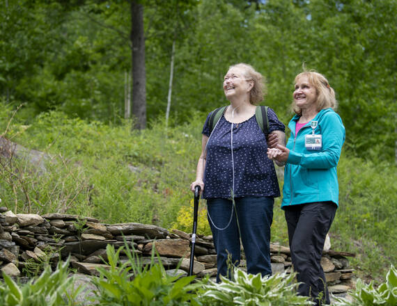 A physical therapist walks arm-in-arm outdoors with a patient using oxygen support and a cane