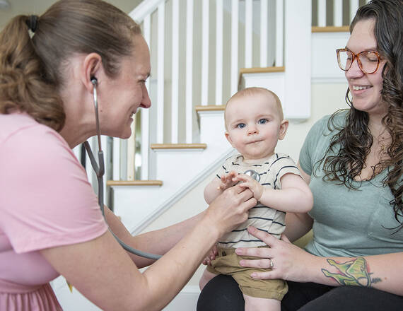 A home health care aide holds a stethoscope to a baby held by its mother.
