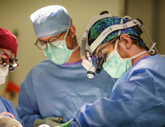 Three surgeons wearing surgical scrubs, masks, and protective eyewear—including headlamps and magnifying loupes—focus intently on a procedure in an operating room.